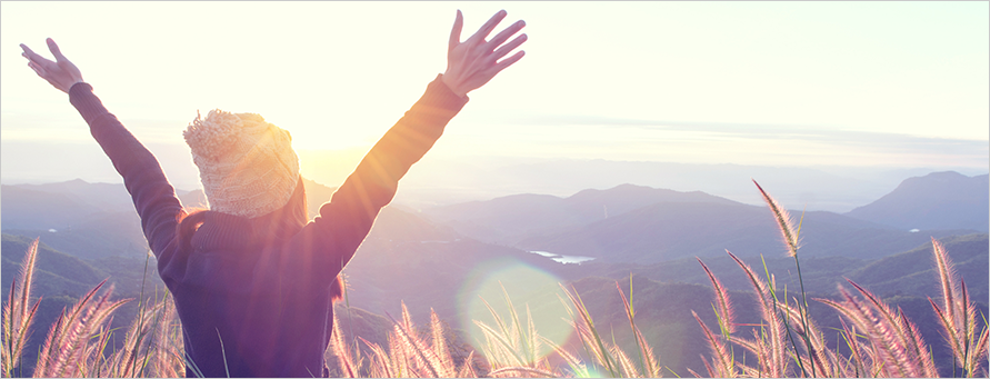 Image of a woman in hat and fleece from the back with her arms stretched out in a Y embracing the sunrise on the horizon from a field atop a large hill, one of the many hills that can be seen to the horizon.